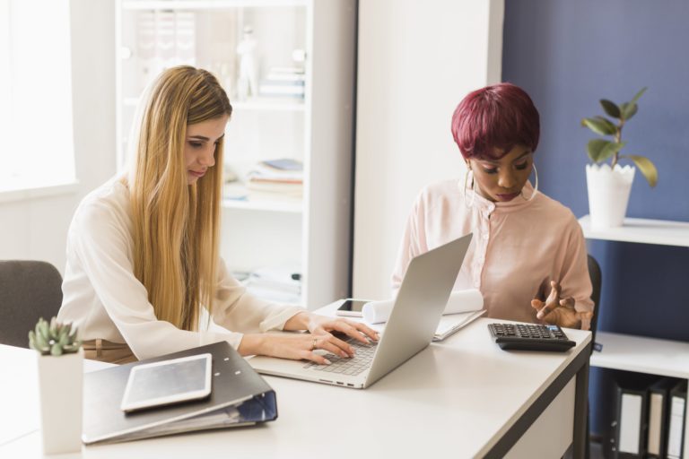 Deux femmes travaillant au bureau; l'une utilise un ordinateur portable, l'autre fait des calculs avec une calculatrice.
