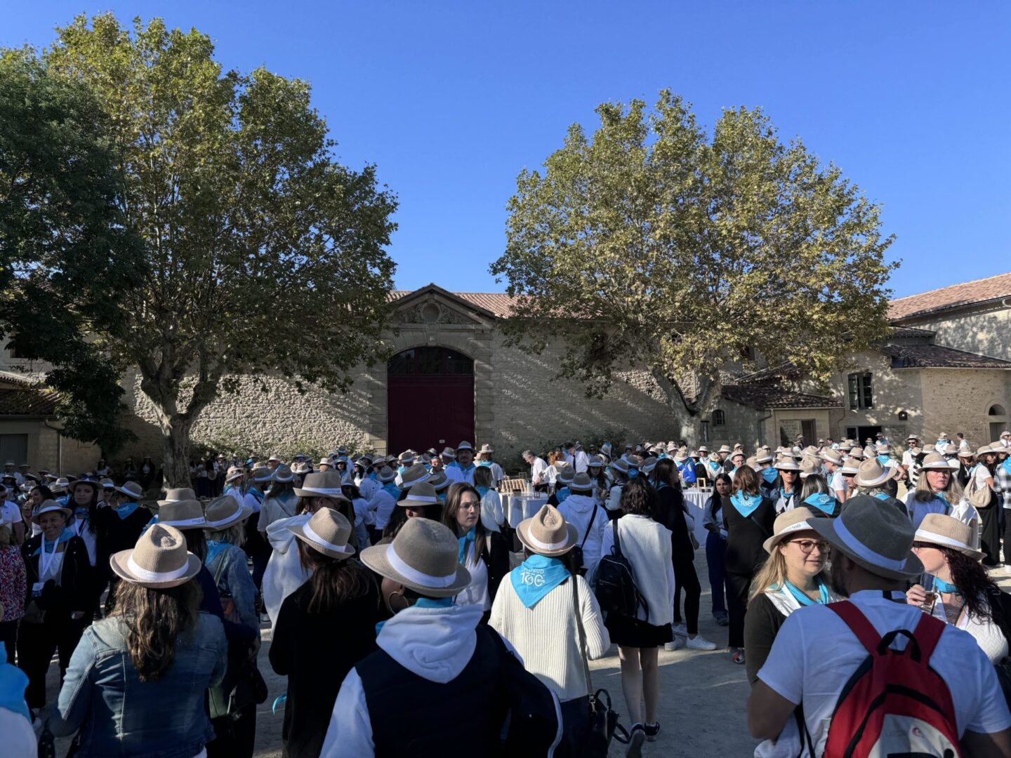 Groupe de personnes portant des chapeaux et foulards bleus, rassemblées devant un bâtiment en pierre sous des arbres.