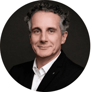 Individu aux cheveux bouclés, portant une veste noire et une chemise blanche, sur un fond sombre.