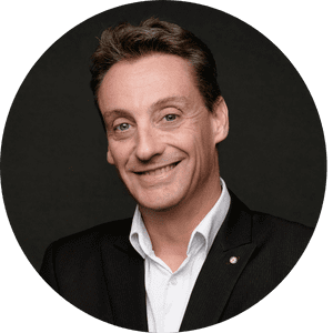 Un homme souriant, en costume sombre et chemise blanche, pose devant un fond noir.