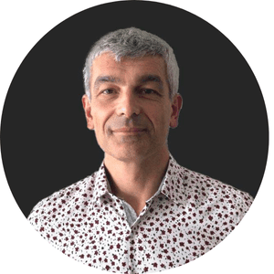 Homme souriant aux cheveux gris, portant une chemise à motifs floraux sur fond noir.