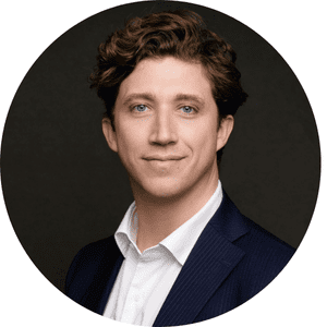 Homme en costume sombre avec chemise blanche, souriant légèrement, sur fond noir.