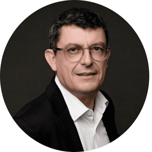 Homme portant des lunettes et une chemise blanche sous une veste noire, souriant légèrement, sur fond sombre.