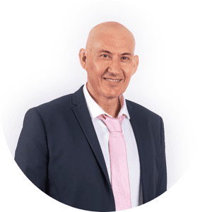 Un homme en costume sombre avec cravate rose pose en souriant, sur un fond blanc.