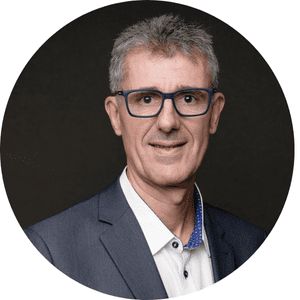 Homme souriant portant des lunettes, vêtu d'un costume sombre et chemise blanche, sur fond noir.