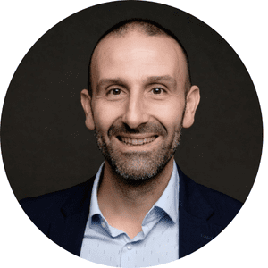 Homme souriant, portant une chemise bleue et veste sombre, sur fond noir.