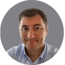Homme aux cheveux bruns, portant une chemise bleue, souriant légèrement devant un fond gris circulaire.