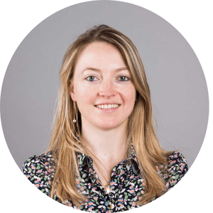 Une femme souriante avec des cheveux blonds, portant une chemise à motifs fleuris, posant devant un fond gris.