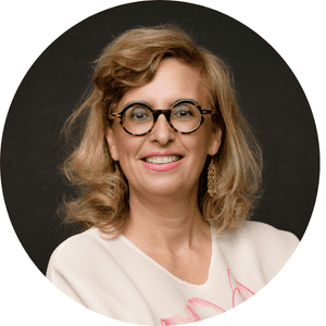 Une femme avec des lunettes et des cheveux blonds sourit, portant un haut clair sur un fond sombre.