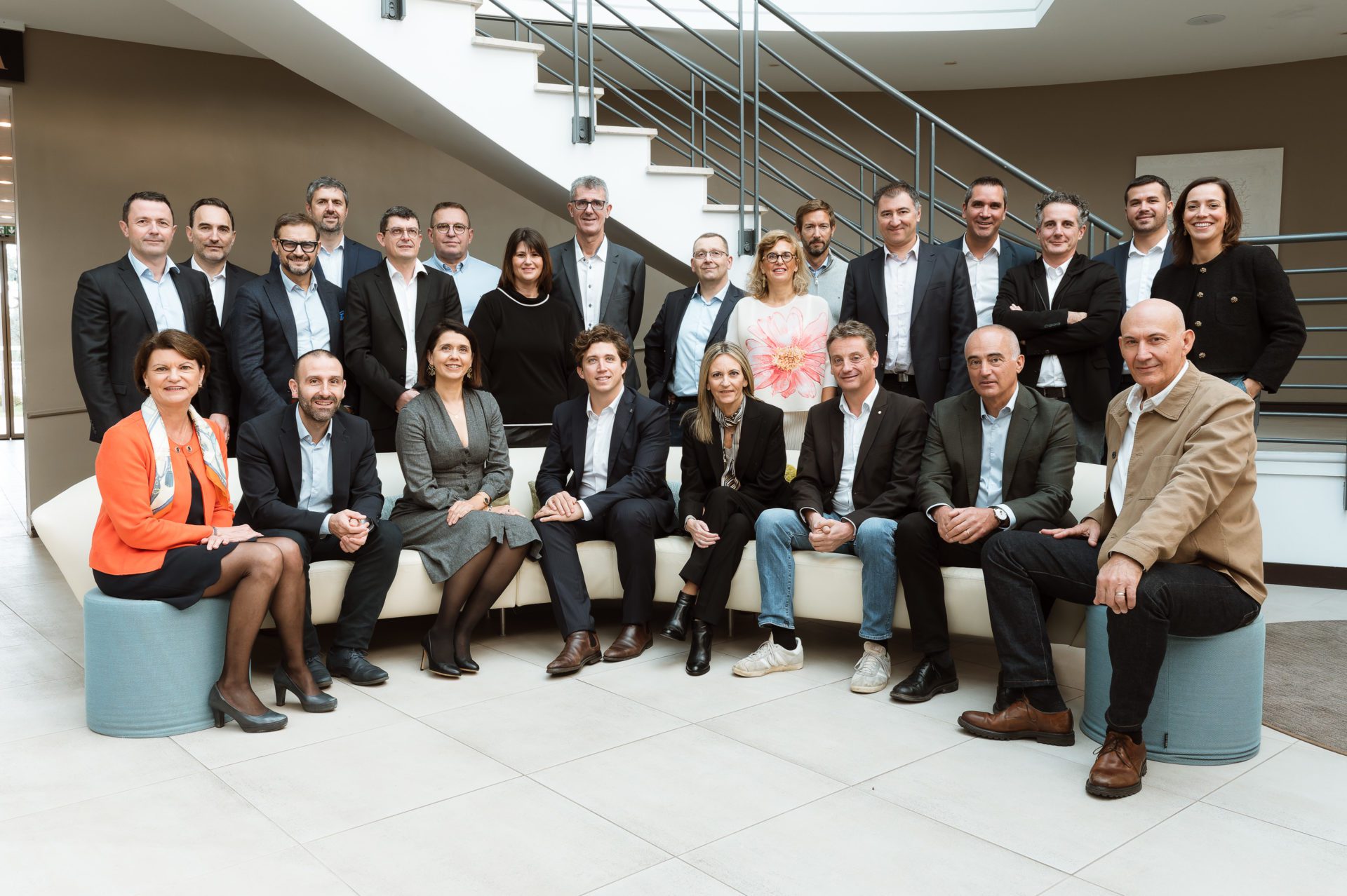 Groupe de 20 personnes en tenue professionnelle posant dans un hall moderne avec escalier en arrière-plan.