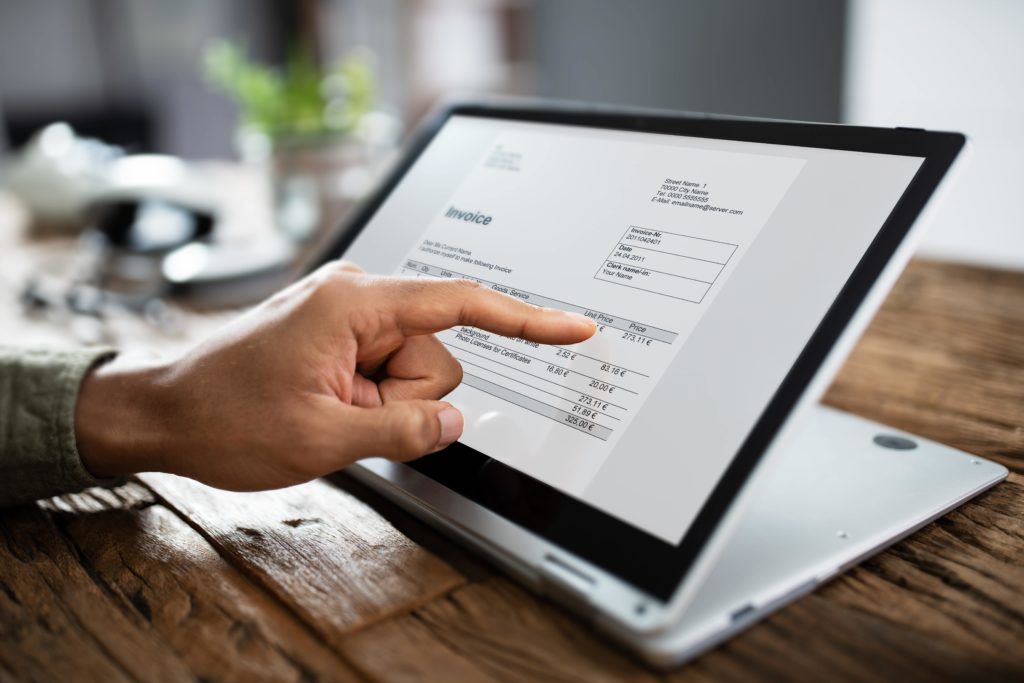 Doigt pointant une facture sur un écran d'ordinateur portable posé sur une table en bois.