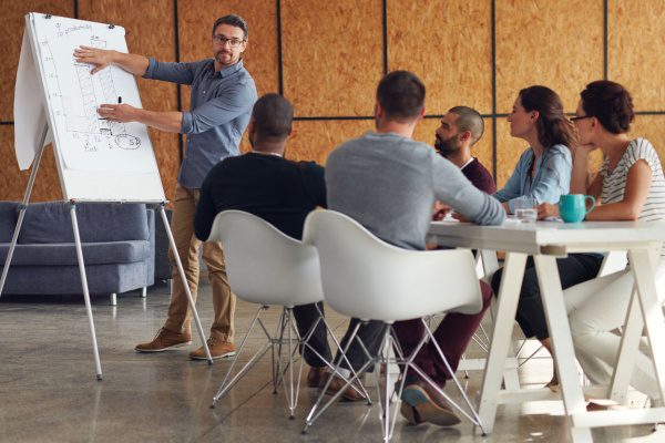 Un homme présente un graphique sur un tableau lors d'une réunion avec cinq personnes attentives autour d'une table.