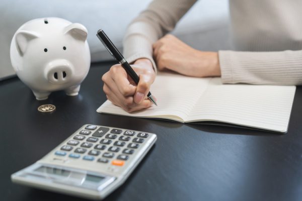 Personne écrivant dans un cahier près d'une calculatrice et d'une tirelire en forme de cochon sur une table noire.