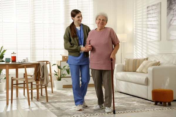 Une professionnelle de santé aide une personne âgée à marcher dans un salon lumineux, utilisant une canne pour se soutenir.