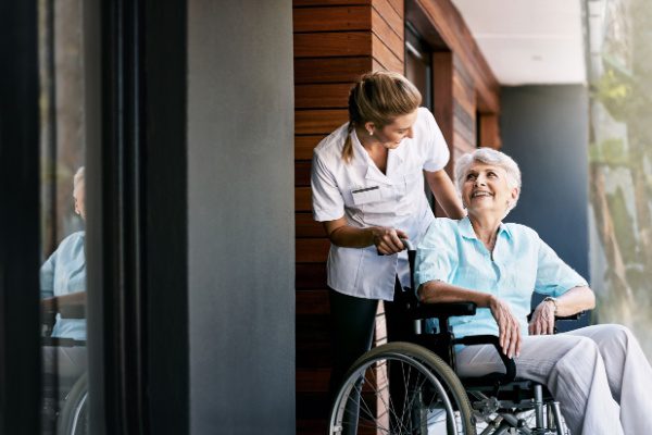 Une soignante souriante aide une personne âgée en fauteuil roulant dans un couloir moderne et lumineux.