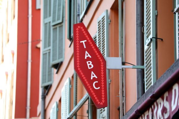 Enseigne rouge de « TABAC » en forme de losange sur un fond de façade orange avec volets verts.