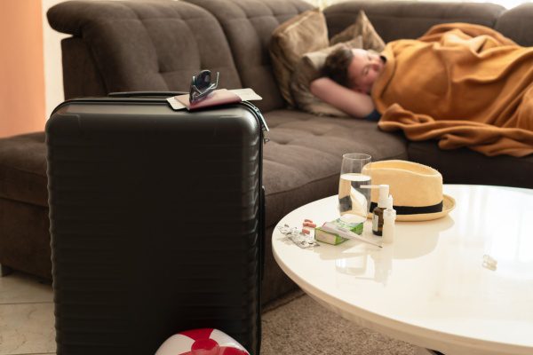 Personne malade enroulée dans une couverture sur un canapé, valise et médicaments sur la table, ambiance de repos.