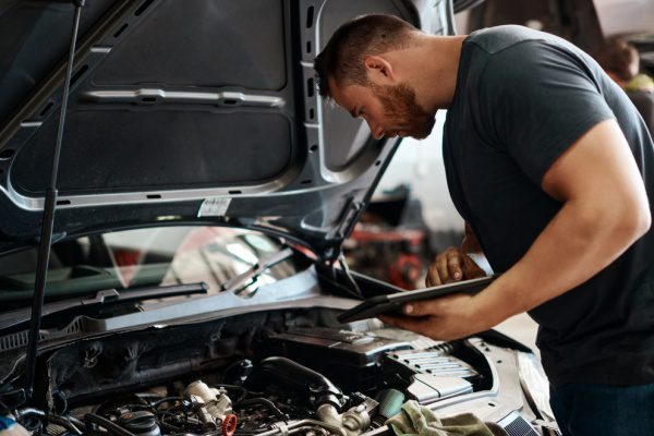 Un homme vérifie le moteur d'une voiture dans un garage, tenant une tablette pour consulter des informations techniques.