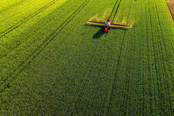 Tracteur rouge pulvérisant un champ vert, vu de haut, avec des lignes parallèles marquées dans le paysage agricole.