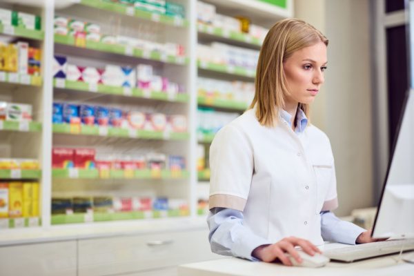 Pharmacienne en blouse blanche utilisant un ordinateur dans une pharmacie, étagères de médicaments en arrière-plan.
