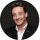 Un homme souriant, en costume sombre et chemise blanche, pose devant un fond noir.