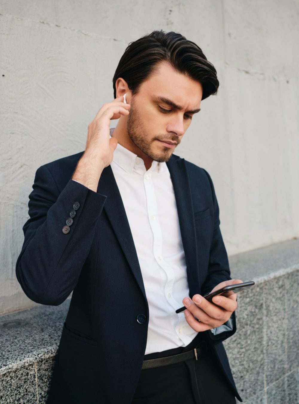 young-beautiful-serious-bearded-brunette-man-in-white-shirt-and-classic-suit-with-wireless-earphones-thoughtfully-using-cellphone-while-standing-near-gray-wall-on-street-min